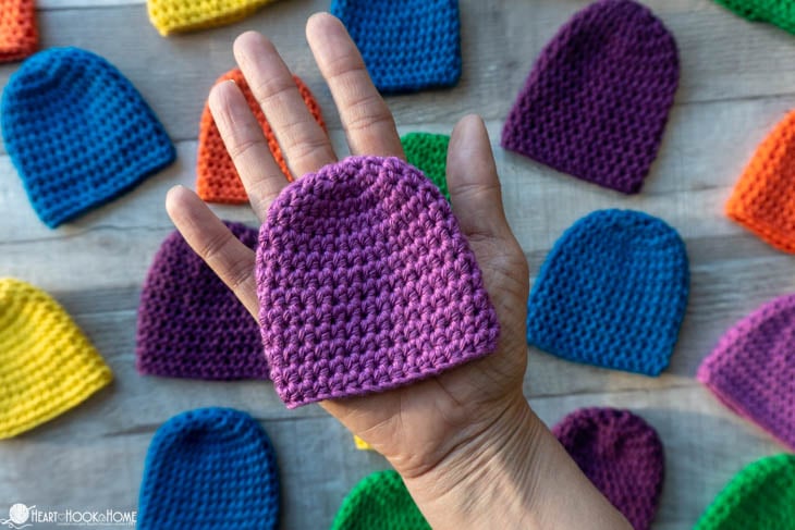 Multiple colors of the Preemie Baby Beanie crochet pattern and a lavender example in an adult\'s hand.