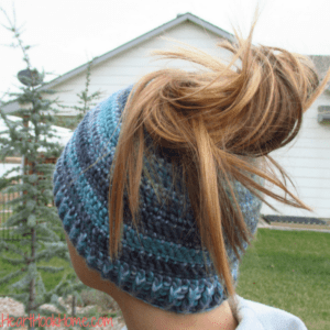Multi-blue colored example of the Messy Mom Bun Beanie crochet pattern with a brunette ponytail coming out of the top.   