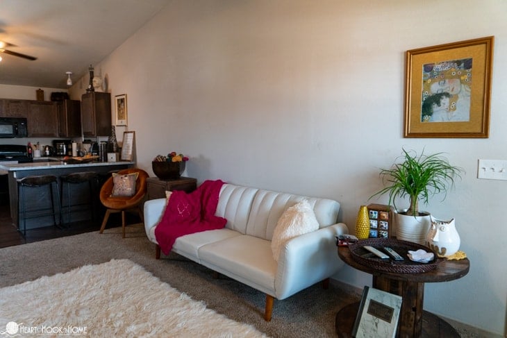White couch with a dragonfruit colored example of a Sangria Sorbet Crochet Blanket pattern draped over it with a white furry rug in the foreground and flanked by end tables with various decorations.  