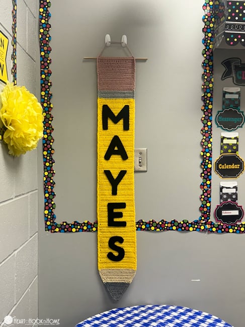 Pink, gray, yellow, and brown example of the Pencil crochet pattern hanging from a wall in a classroom setting with the name Mayes on it in black.