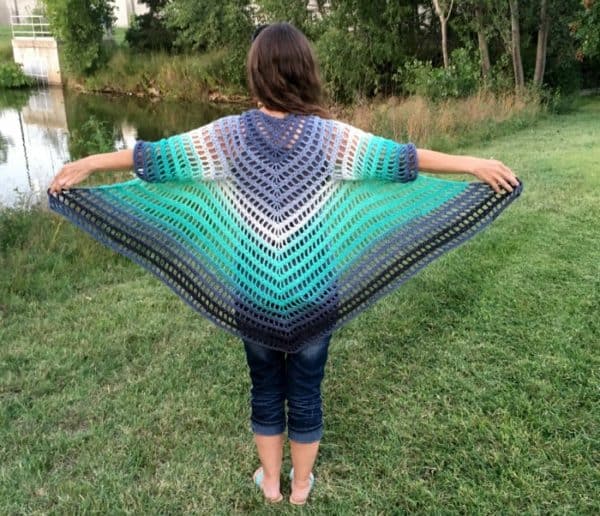Back of a blue, white, and gray example of the 12/14/16 size of the It's Shawl Good crochet pattern on a teen model wearing jeans, outstretched arms, and standing in a grassy setting.