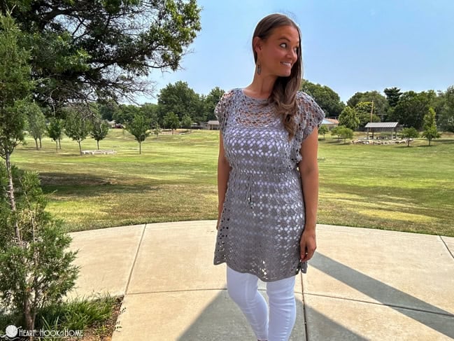 Smiling female model wearing an example of the Shell Yeah! Tunic crochet pattern in Comfy Fingering yarn in the “Whisker” colorway in a park setting.