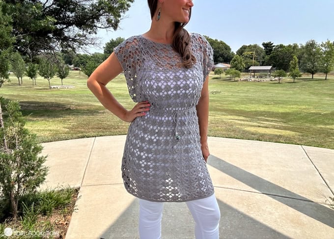 Smiling female model wearing an example of the Shell Yeah! Tunic crochet pattern in Comfy Fingering yarn in the “Whisker” colorway in a park setting.