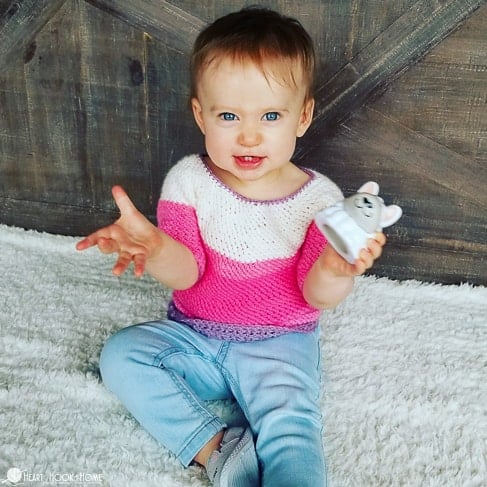 Pink, white, and purple example of the Shanti Sweater for Little Kids crochet pattern on a smiling, female child wearing jeans and sitting on a white rug.