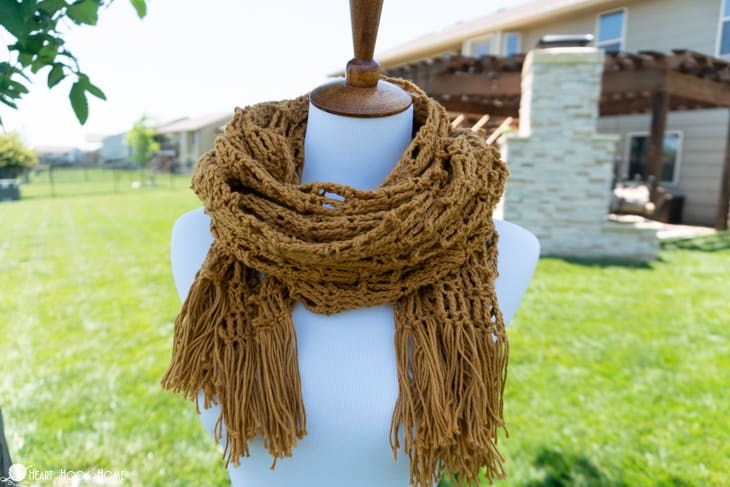 Gold example of the Let It Bee Wrap crochet pattern wrapped around the neck of a white dress form in a backyard setting.