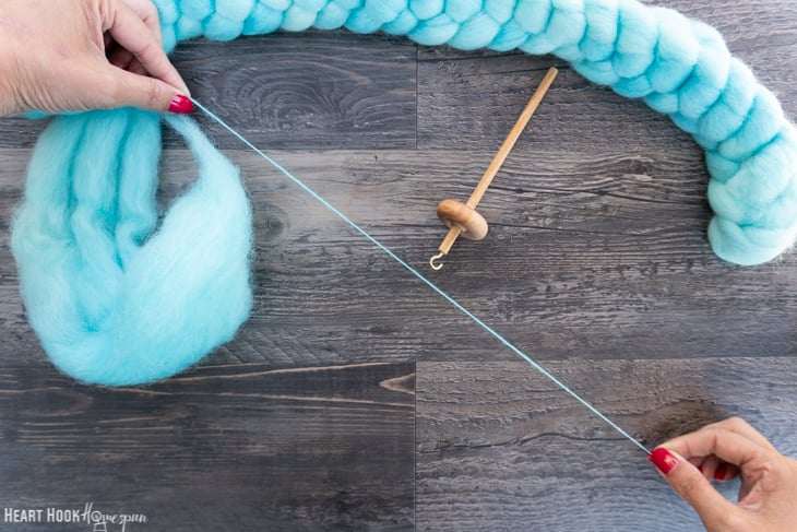 Braid of blue roving yarn and a wood drop spindle with two hands carefully drafting some yarn.