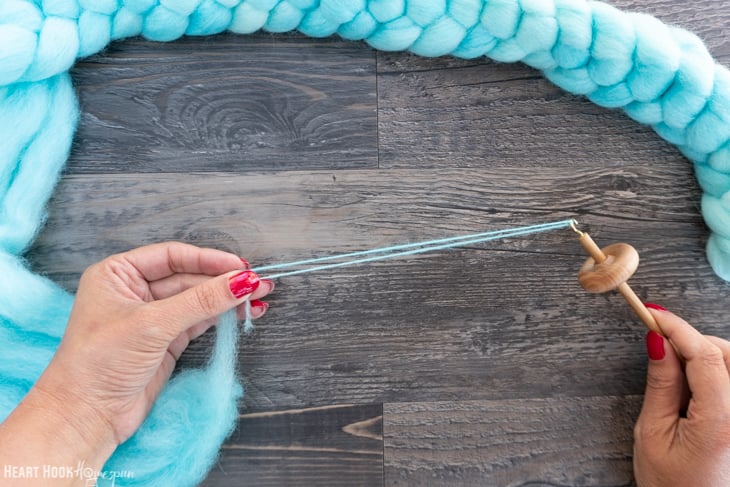 Braid of blue roving yarn and a wood drop spindle with two hands twisting until yarn starts to form.