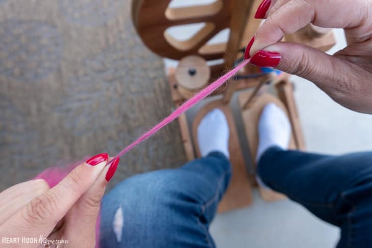 Example of a slub in pink, handspun yarn with a spinning wheel and a pair of legs in the background.