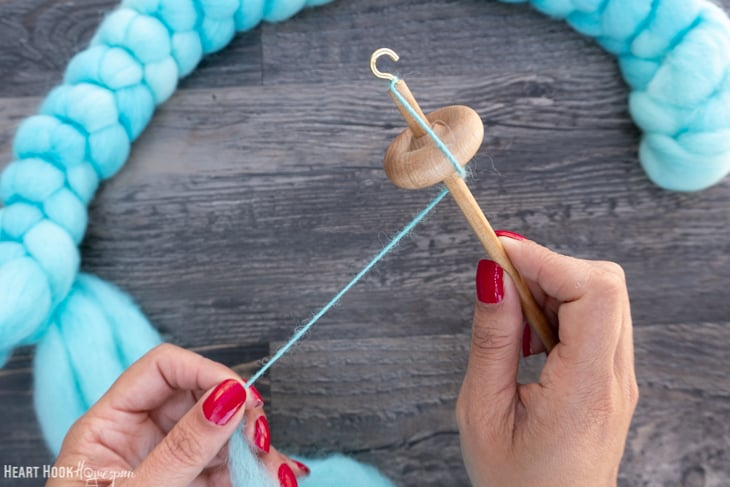 Braid of blue roving yarn and a wood drop spindle with one hand twisting the yarn and the other holding the drop spindle.