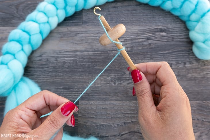 Braid of blue roving yarn and a wood drop spindle with one hand twisting the yarn and the other holding the drop spindle.