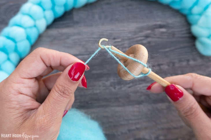 Braid of blue roving yarn and a wood drop spindle with one hand twisting the yarn and the other holding the drop spindle.