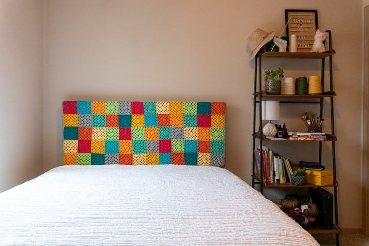 Plain white bed with an example of how to yarn bomb a headboard crochet pattern in various colors.  Nightstand next to it has books, a hat, yarn cones, a plant, paintbrushes in a bowl, and a basket of yarn.