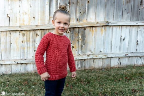 Pink version of the child size Color Block Sweater crochet pattern on a smiling female child wearing jeans in front of a wood fence standing on grass.