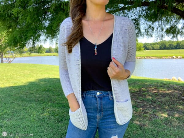 Gray version with pockets of the Rose Cardi crochet pattern on a female model wearing a dark shirt and jeans in a park setting.