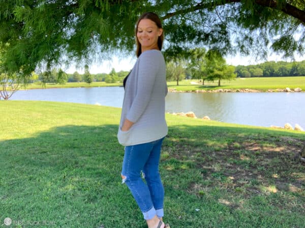 Smiling female model wearing jeans with her hand in the pocket of a gray version of the Rose Cardi crochet pattern that she is wearing in a park setting.