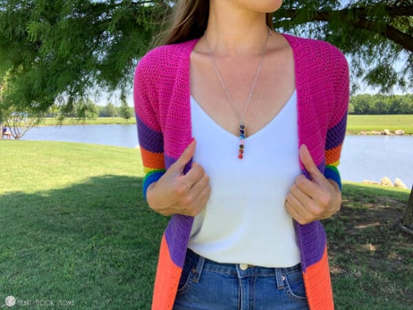 Female model wearing a rainbow version of the Rose Cardi crochet pattern over a white top and jeans in a park setting near a pond.
