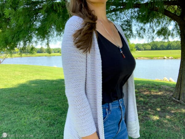 Female model wearing a gray version of the Rose Cardi crochet pattern over a dark top and jeans in a park setting near a pond.  The fitted sleeves are the prominent aspect.