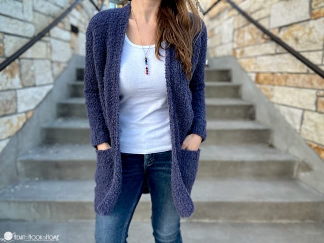 Example of the Bernadette Boucle Cardi crochet pattern on a female model with a white undershirt and jeans standing on concrete stairs.