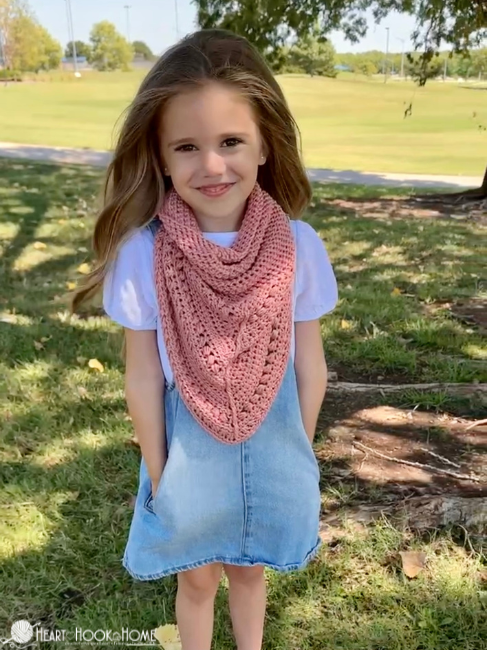 Smiling female child wearing a light blue denim dress and a Lion Brand Coboo yarn in the “mauve” colorway example of the Frankie Cowl crochet pattern in a park setting.