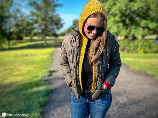 Gold colorway example of the Layering Hooded Vest crochet pattern on a smiling female model wearing a brown jacket, jeans, and a black undershirt in a park setting.