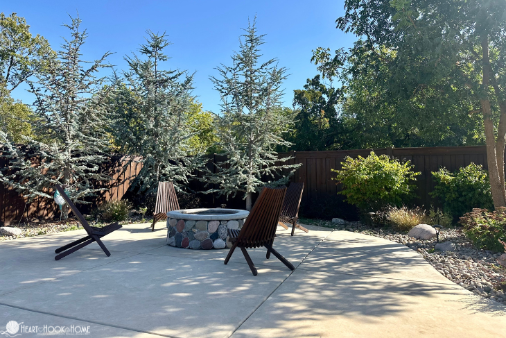 Stone outdoor fire pit on a concrete patio surrounded by various trees and wood lounge chairs.