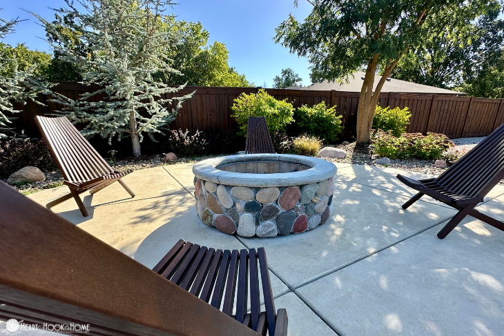 Stone outdoor fire pit on a concrete patio surrounded by various trees and wood lounge chairs.