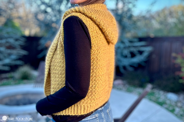 Gold example of the back of a Layering Hooded Vest crochet pattern on a female model wearing a black, long sleeved shirt and jeans in a backyard setting.