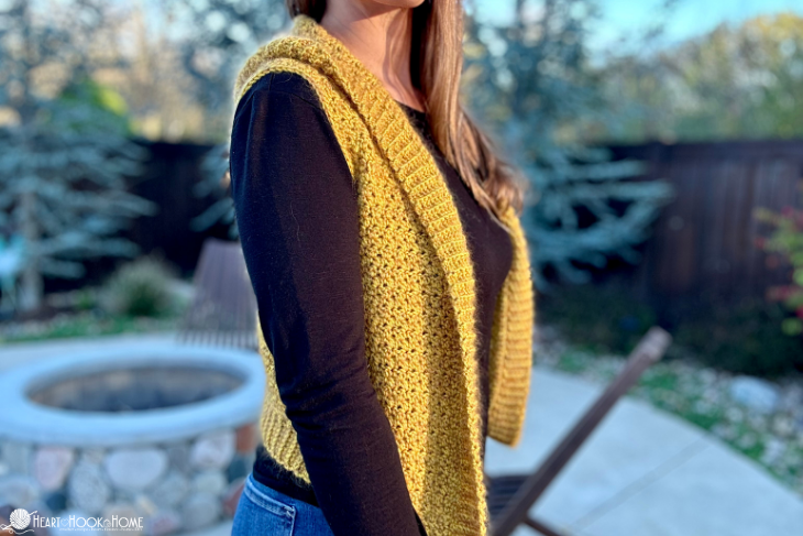 Gold example of the front of the Layering Hooded Vest crochet pattern on a female model wearing a black, long sleeved shirt and jeans in a backyard setting.
